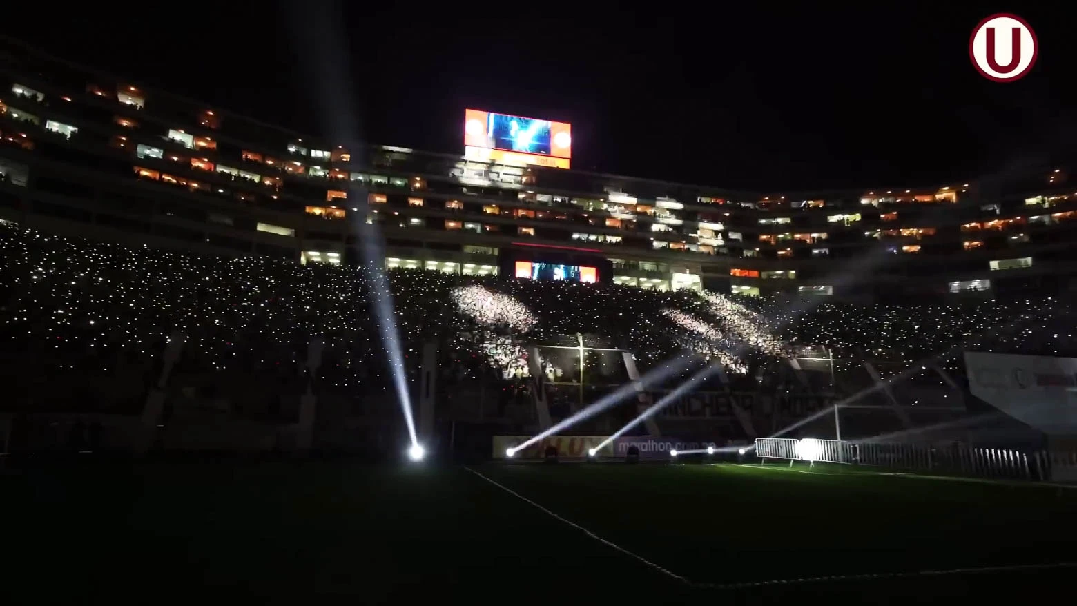 Vista nocturna del Estadio Monumental de Lima durante un partido de fútbol, con el campo iluminado por luces de cabezales móviles y las gradas llenas de aficionados que usan pulseras LED DMX inalámbricas sincronizadas con el juego - Iluminación espectacular en Noche Crema 2024 con Scratch Perú - scratch perú.