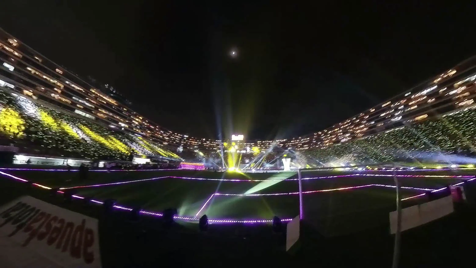 Vista desde el exterior de un estadio de fútbol con un túnel triangular de tubos LED en la entrada, que invita a los visitantes a pasar por él y disfrutar de una experiencia sensorial de luz y sonido - Iluminación espectacular en Noche Crema 2024 con Scratch Perú - scratch perú.