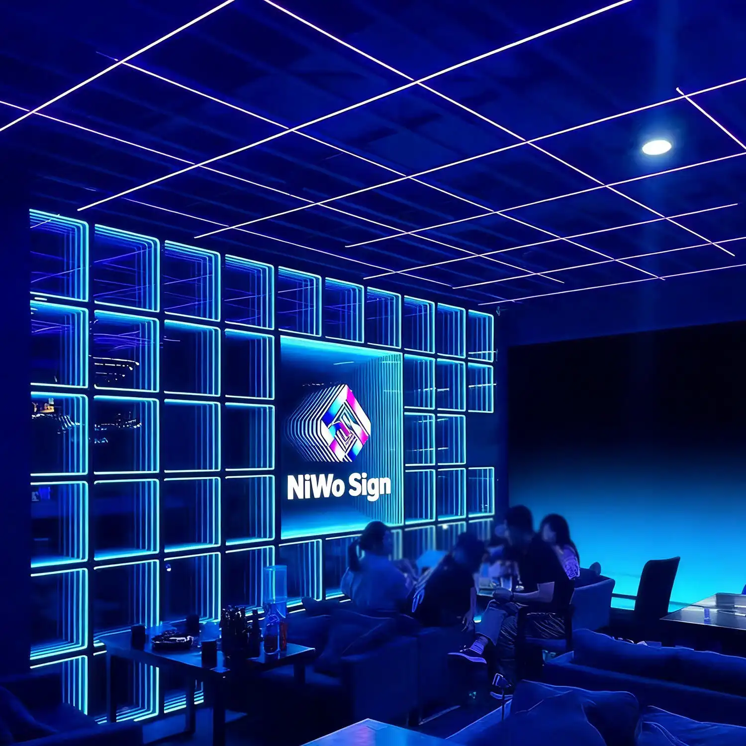 Pared LED Espejo Infinito 3D en un lounge nocturno Pared de paneles LED Espejo Infinito 3D en tono azul intenso con el logo NiWo Sign, ambientando un lounge nocturno con personas sentadas.