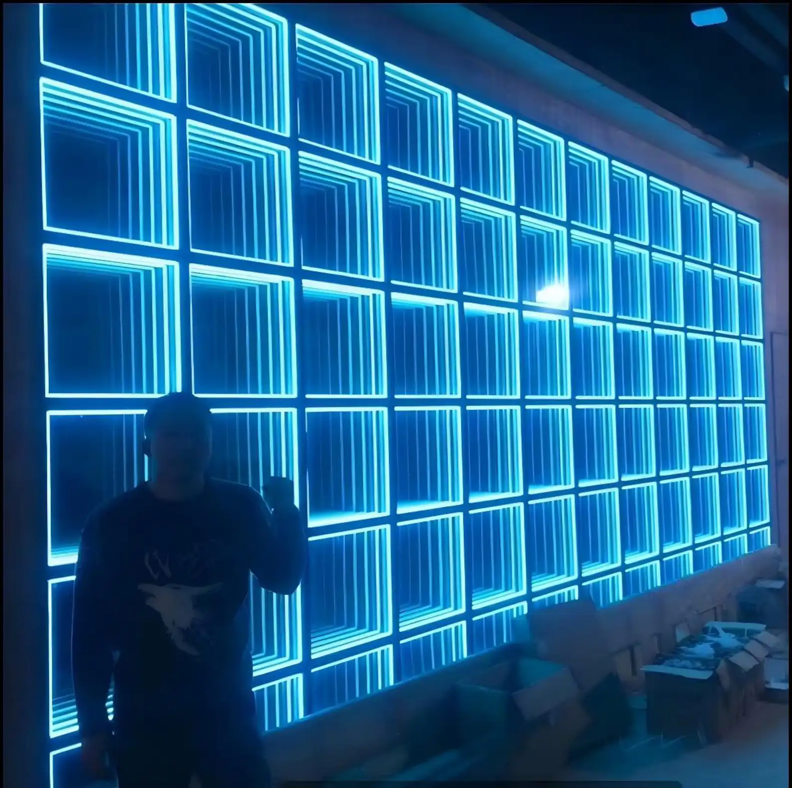 Pared LED Espejo Infinito 3D de gran formato en tono azul Pared de gran formato revestida con paneles LED Espejo Infinito 3D en tono azul, con una persona de pie que permite apreciar la escala de la instalación.