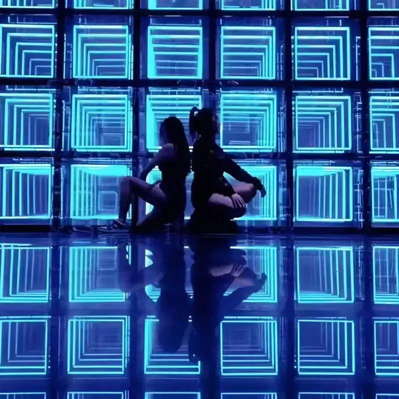 Bailarinas en silueta frente a una pared LED Espejo Infinito 3D azul Dos bailarinas en silueta posando frente a una pared de paneles LED Espejo Infinito 3D en tono azul turquesa, con su reflejo proyectado en el piso.