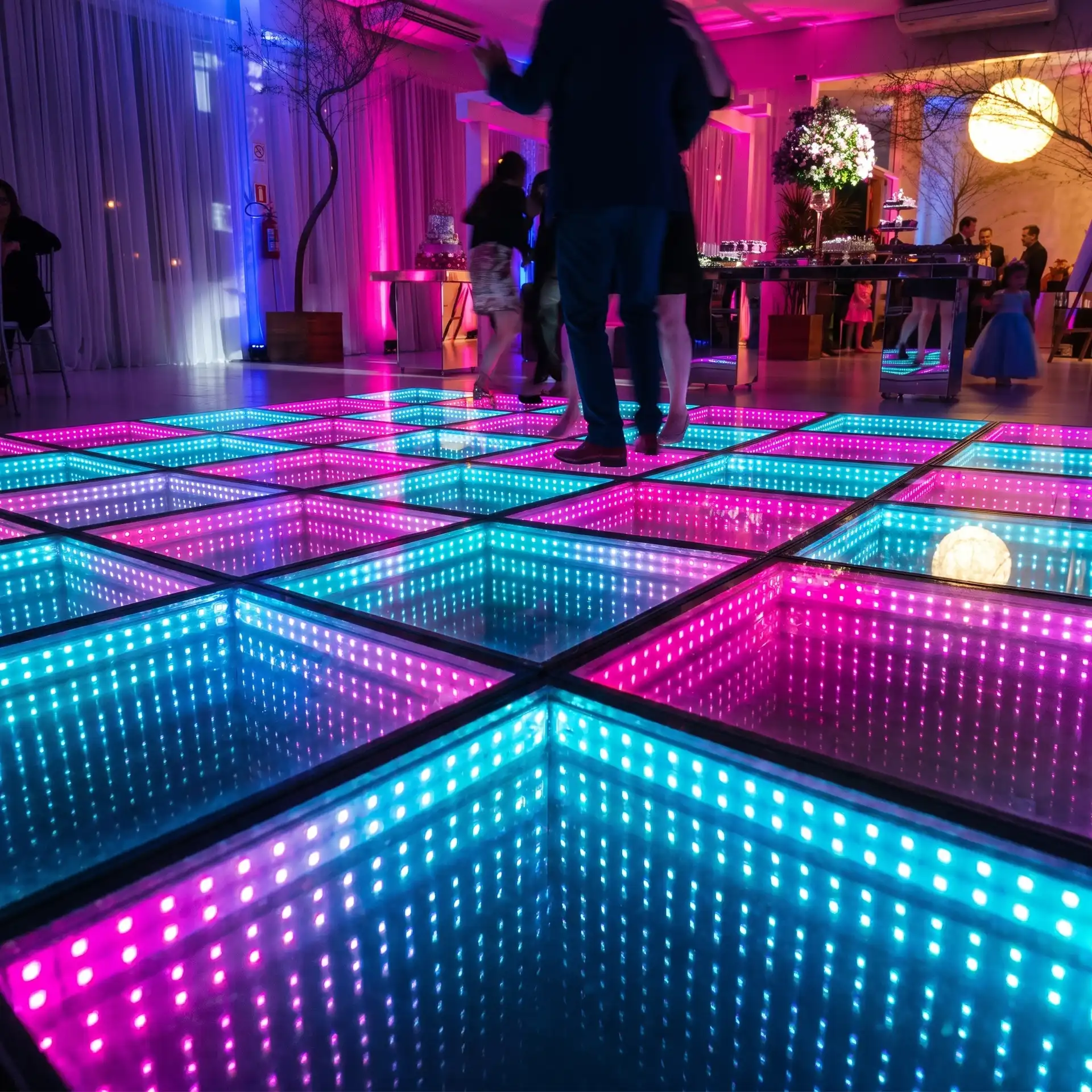 Pareja bailando en pista LED 3D Infinity Mirror rosa y turquesa durante una fiesta Invitados bailando sobre la pista de baile LED 3D Infinity Mirror en tonos rosa y turquesa, con mesa de postres y decoración floral al fondo.