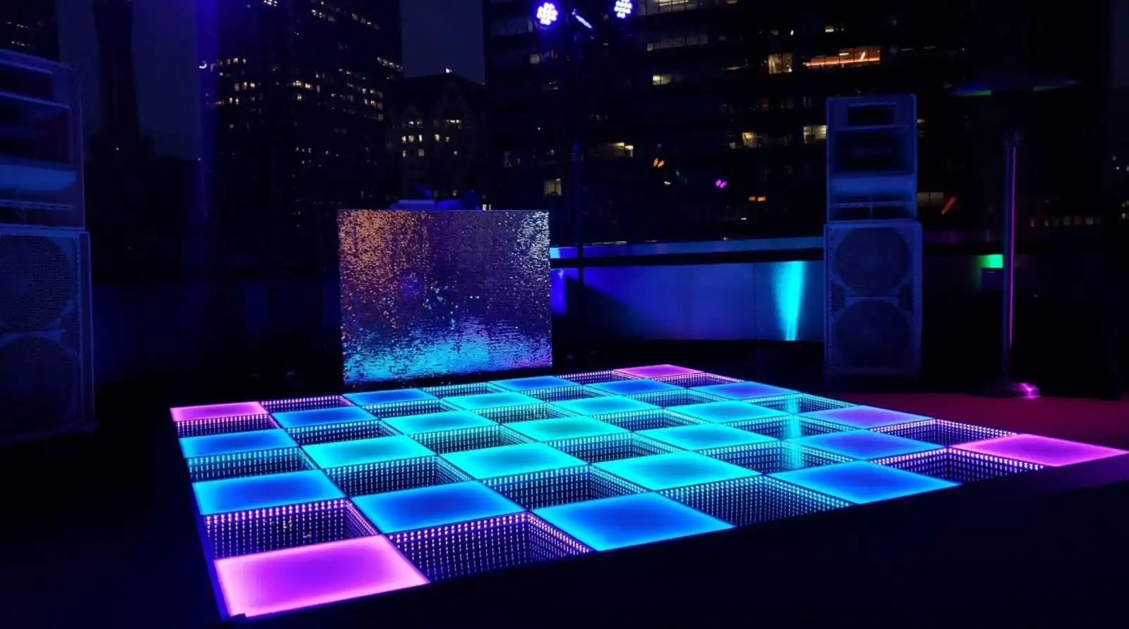 Pista LED 3D Infinity Mirror azul y púrpura en una fiesta en azotea Pista de baile LED 3D Infinity Mirror en tonos azul y púrpura instalada en una terraza nocturna con vista al skyline de la ciudad.