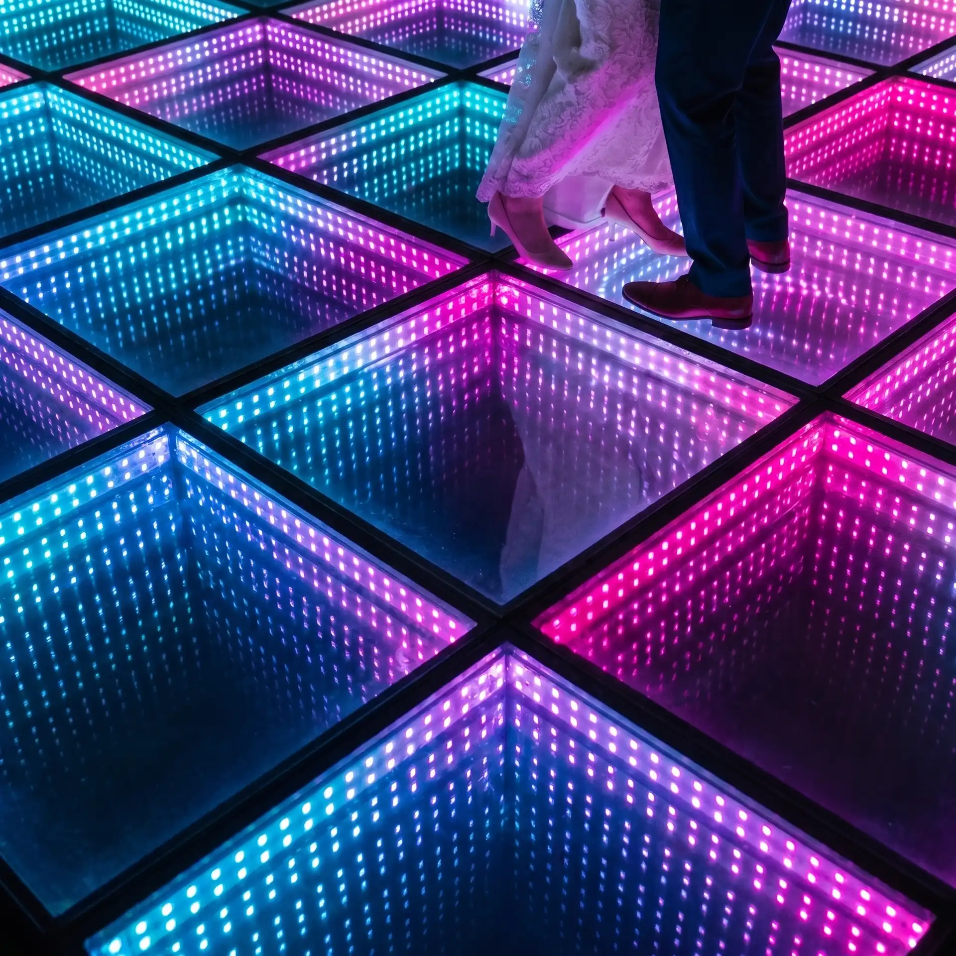 Novios bailando sobre pista LED 3D Infinity Mirror en una boda Detalle de los pies de los novios bailando sobre la pista de baile LED 3D Infinity Mirror en tonos rosa, púrpura y turquesa durante la recepción de la boda.