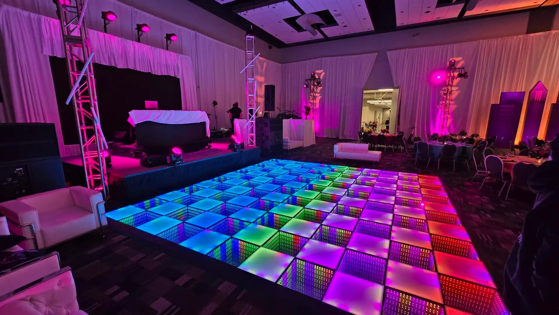 Pista LED 3D Infinity Mirror multicolor en un salón con cabina de DJ Pista de baile LED 3D Infinity Mirror en tonos rosa, azul y verde en un salón de eventos con cabina de DJ y mesas para invitados.