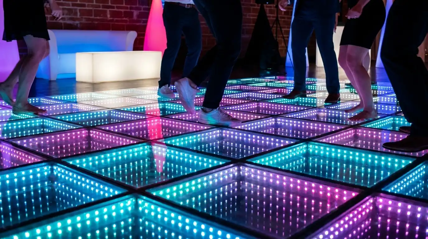 Invitados bailando en la pista LED 3D Infinity Mirror junto a una pared de ladrillo Invitados descalzos y con tacones bailando sobre la pista de baile LED 3D Infinity Mirror en tonos rosa, púrpura y turquesa, junto a una pared de ladrillo expuesto.