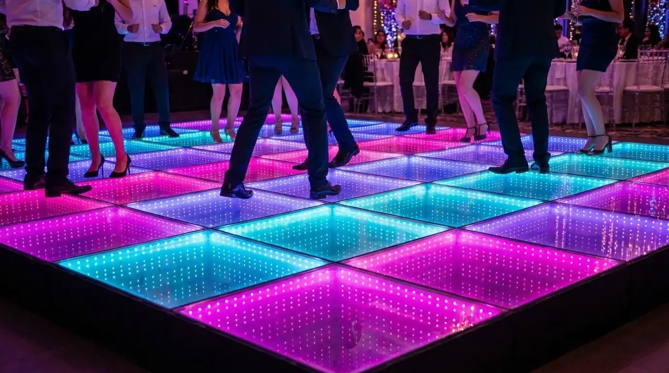 Invitados bailando sobre la pista LED 3D Infinity Mirror en una celebración de fin de año Invitados bailando sobre la pista de baile LED 3D Infinity Mirror en tonos rosa, turquesa y azul en un salón decorado para una celebración de fin de año.