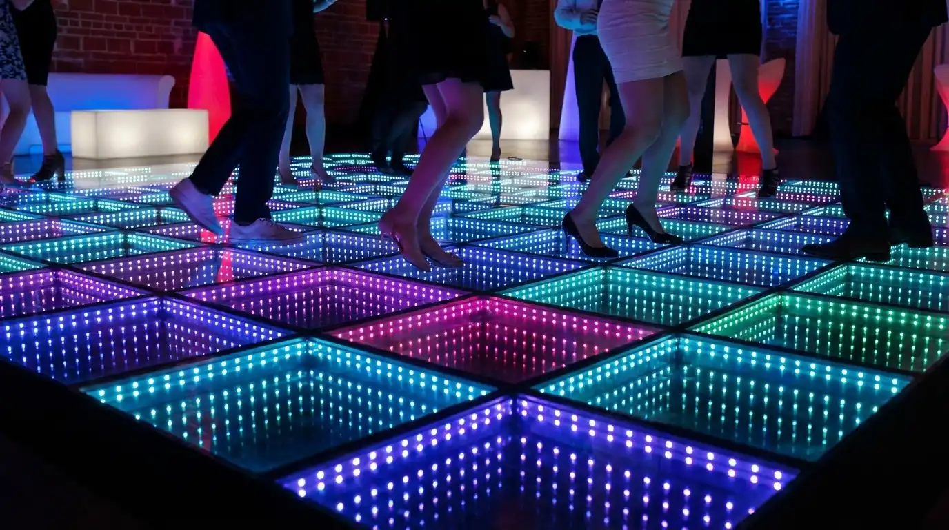 Invitados bailando sobre la pista LED 3D Infinity Mirror multicolor junto a mobiliario lounge Invitados bailando sobre la pista de baile LED 3D Infinity Mirror multicolor junto a una pared de ladrillo y mobiliario lounge iluminado.