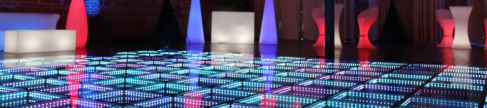 Pista LED 3D Infinity Mirror azul junto a mobiliario iluminado en una terraza Pista de baile LED 3D Infinity Mirror en tono azul junto a mobiliario lounge iluminado en tonos blanco, rojo y azul, en una terraza con pared de ladrillo.