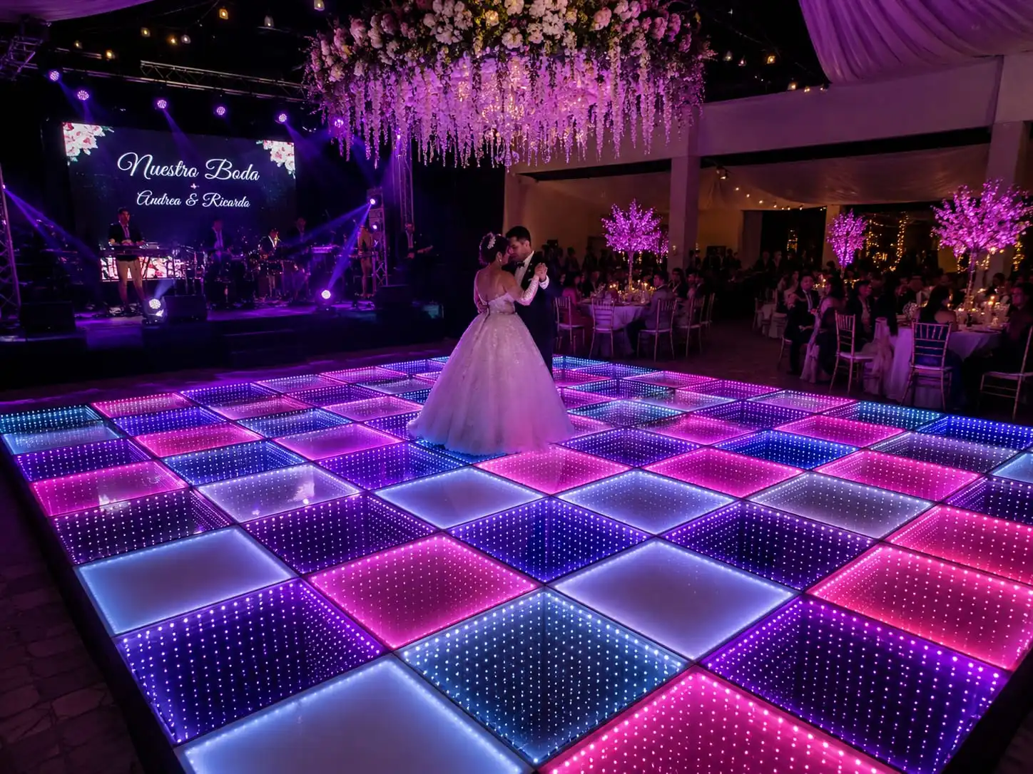 Primer baile de los novios sobre la pista LED 3D Infinity Mirror Los novios Andrea y Ricardo bailando su primer baile sobre la pista de baile LED 3D Infinity Mirror en tonos rosa y púrpura, con banda en vivo y decoración floral.