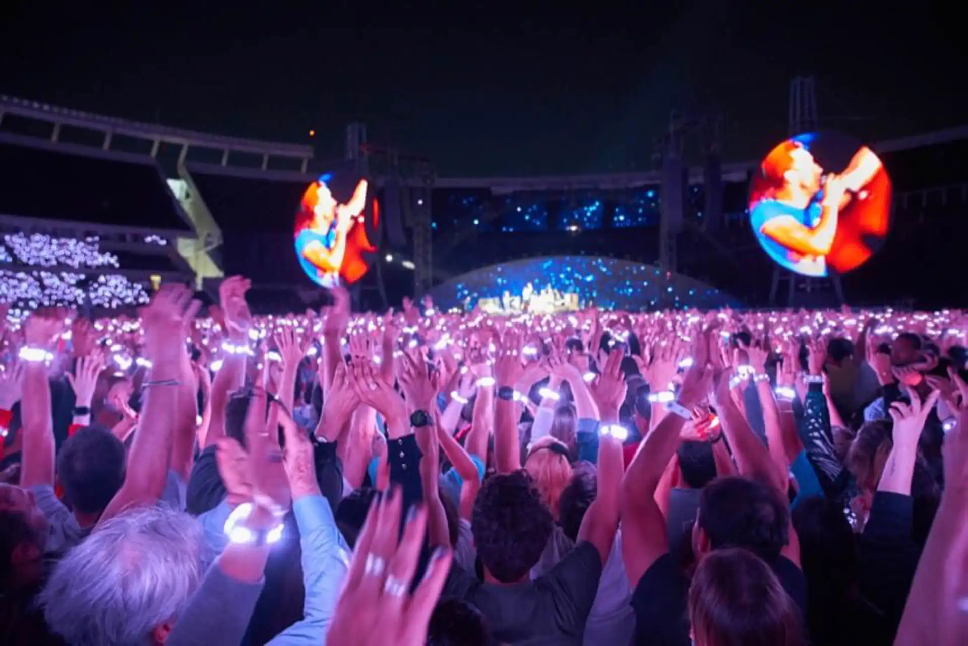 Multitud levantando las manos con pulseras LED blancas sincronizadas, creando un efecto visual impresionante en un concierto.